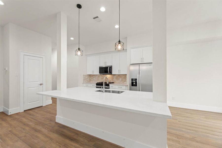 This modern kitchen features a large island with a sleek white countertop and built-in sink. It includes stainless steel appliances, bright pendant lighting, and ample cabinet space, all set against a backdrop of light flooring.