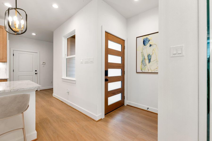 Welcoming entryway with a striking modern wood front door featuring frosted glass panels, allowing natural light to flow inside. Warm wood floors, recessed lighting, and crisp white walls create a bright, open, and contemporary atmosphere that sets the tone for the home.