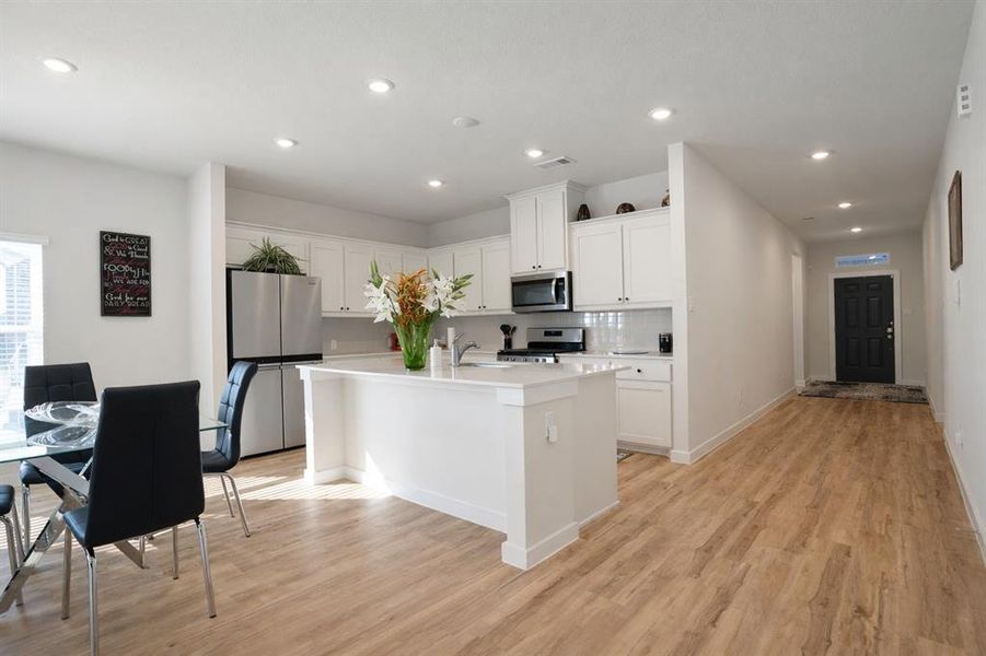 Modern open-concept living area featuring wood-finish flooring, recessed lighting, a kitchen island with a white countertop, white cabinetry, and stainless steel appliances