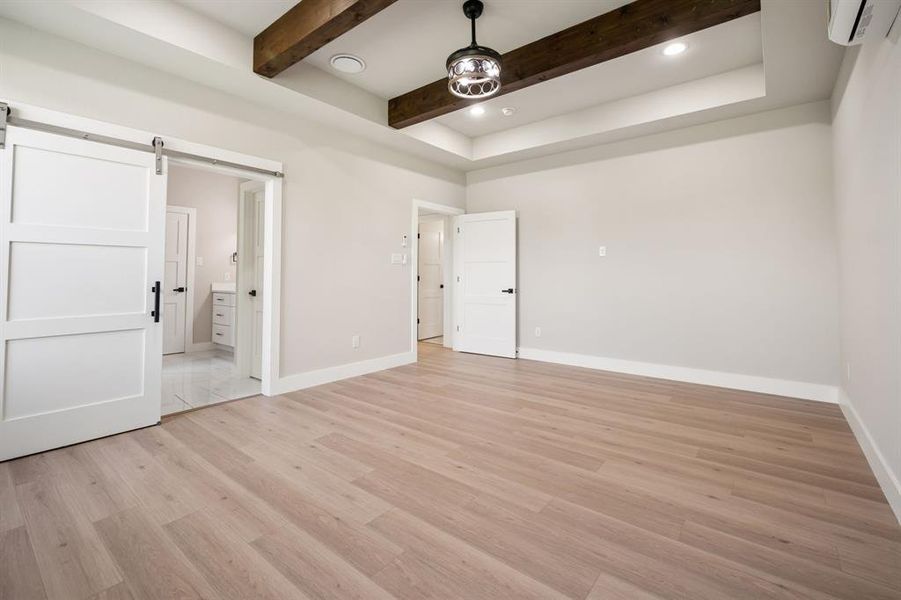 First floor primary bedroom with barn door entry to bath, recessed lighting, ensuite bathroom, light wood finished floors, and beam ceiling
