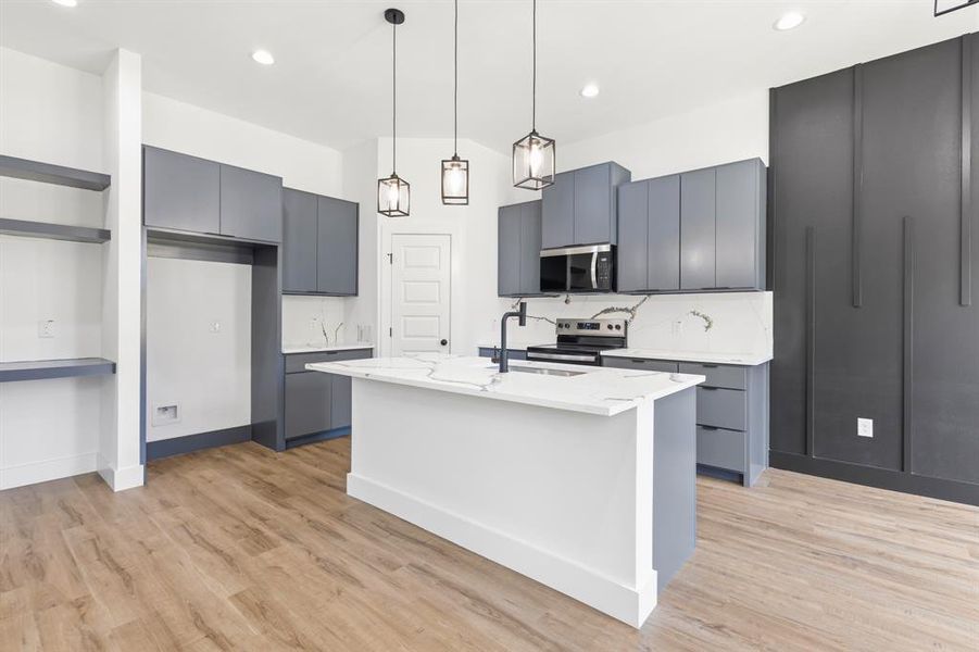 Modern kitchen featuring a central island with a waterfall-edge countertop, grey cabinetry, stainless steel appliances, a paneled accent wall, and wood-finish flooring Modern kitchen featuring a central island with a waterfall-edge countertop, grey cabinetry, stainless steel appliances, a paneled accent wall, and wood-finish flooring