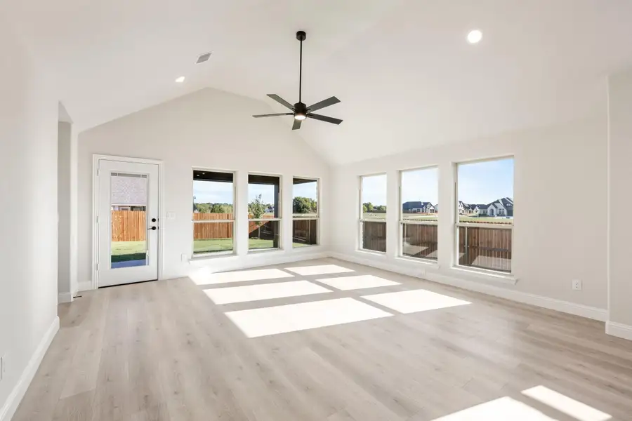 Open living area with vaulted ceiling, ceiling fan, light wood floors, and large windows with backyard views