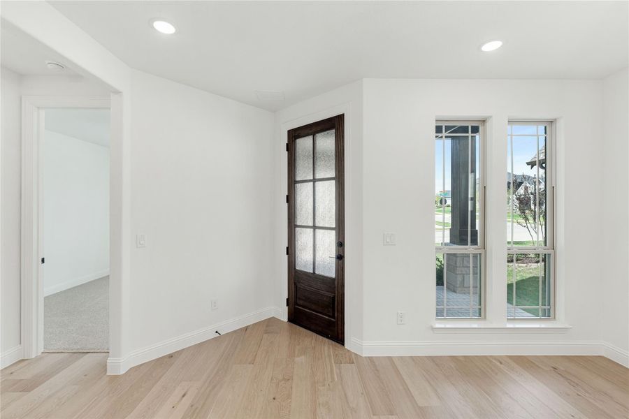 Empty room with light hardwood floors, white walls, dark wood glass-panel door, and double windows with outdoor view. Empty room with light hardwood floors, white walls, dark wood glass-panel door, and double windows with outdoor view.