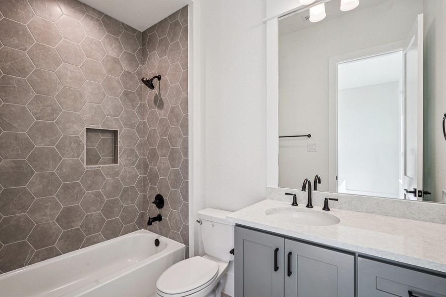 Modern bathroom featuring a bathtub with stylish hexagonal gray tiles, a recessed shelf, and sleek black fixtures. The vanity includes a large mirror, white countertop, and contemporary black faucet, complemented by a soft gray cabinet.