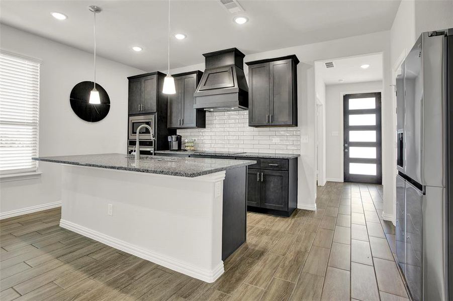 Kitchen featuring tasteful backsplash, premium range hood, an island with sink, stainless steel appliances, and decorative light fixtures