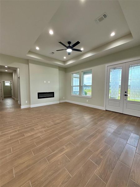 This spacious living area features stylish wood-look tile flooring, a modern recessed ceiling with a contemporary fan, and a sleek built-in fireplace. Natural light floods through large windows and glass doors, offering a view of the outdoors and easy access to the patio.