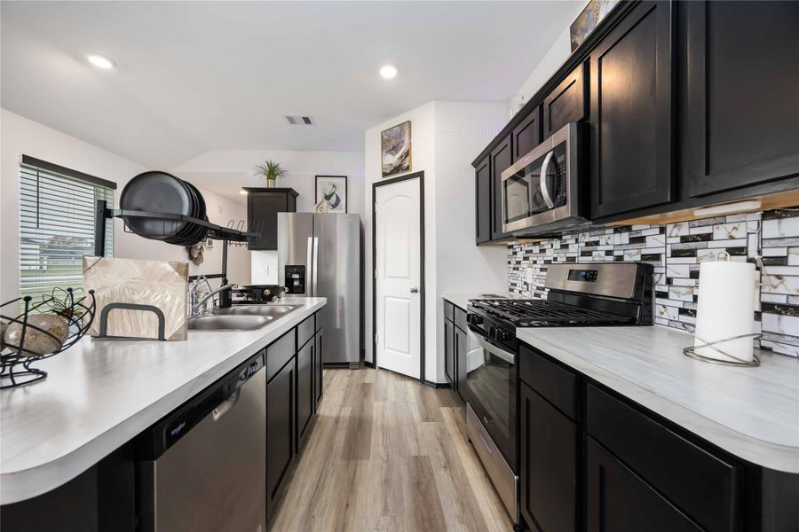 Modern kitchen with sleek black cabinets, stainless steel appliances, and a stylish mosaic backsplash. Features a spacious layout with ample counter space and a large window for natural light.