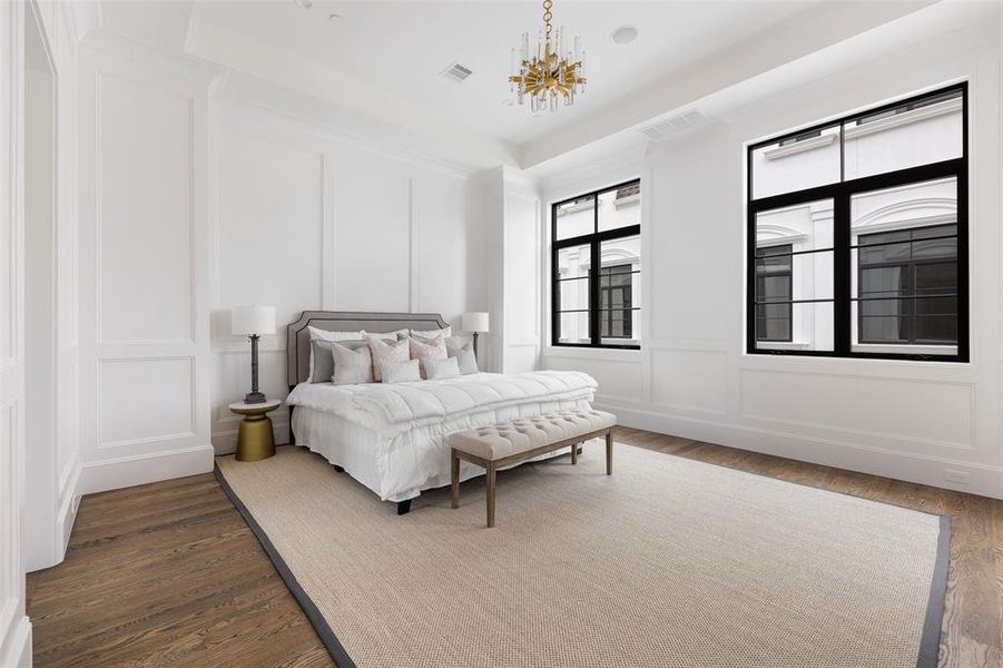 The luxurious master bedroom boasts soaring 12' tray ceiling with elegant floor to ceiling paneling. A designer statement chandelier adds a bit of glam and contrasts the bright millwork and dark casement windows. Image from previously completed home in the community.