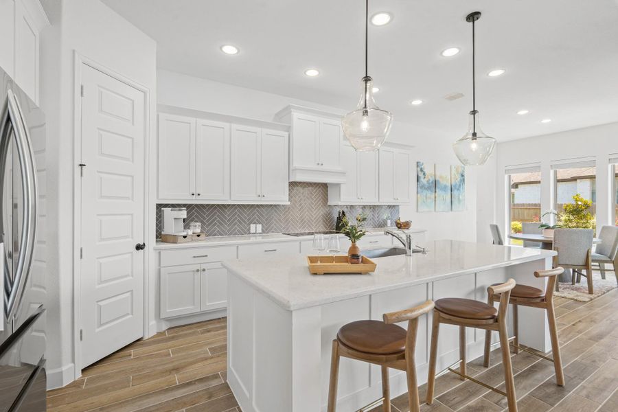 This modern kitchen features sleek white cabinetry, a spacious island with bar seating, and stylish pendant lighting. It opens to a bright dining area with large windows, offering plenty of natural light and a view of the backyard.
