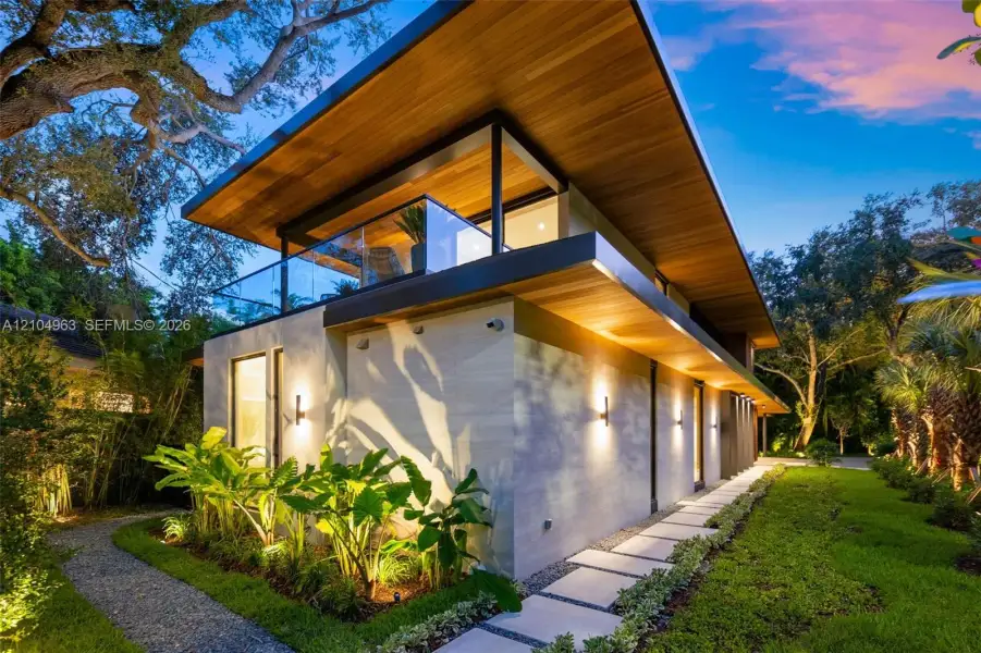 Modern architectural exterior featuring a wood-paneled cantilevered roof, stone-clad facade, glass-paneled balcony railing, and paver stone walkway