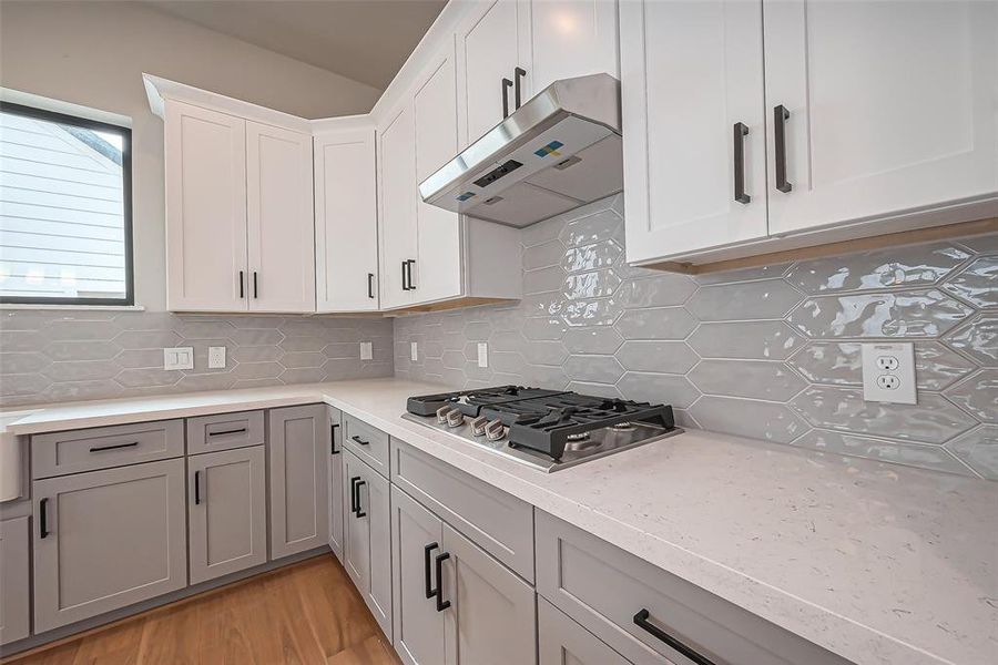 This modern kitchen boasts sleek stainless steel appliances, light wood flooring, and a spacious central island. The stylish white and gray cabinetry is enhanced by contemporary pendant lighting and a neutral backsplash.