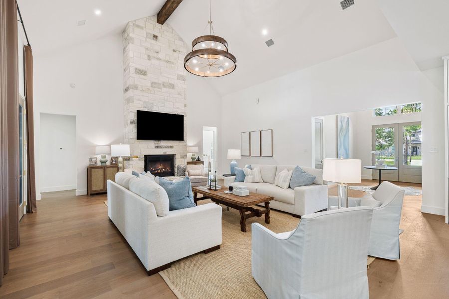 Grand open living space highlighted by a striking oversized chandelier and soaring ceilings that immediately draw your eye up. Two sets of French doors line the back wall, filling the room with natural light and providing direct access to the outdoor living area. The floor-to-ceiling stone fireplace adds texture and scale, while the wide-plank wood flooring carries seamlessly into the kitchen.