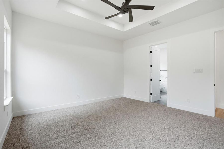Spacious room featuring neutral carpeting, white walls, a tray ceiling with a ceiling fan, and a window with white trim