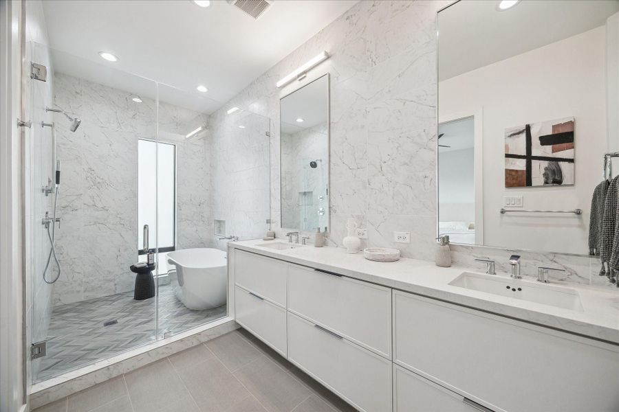 Spa-inspired primary bath wrapped in marble-look tile, featuring dual vanities, expansive mirrors, and a freestanding soaking tub positioned beside a glass-enclosed shower. Elegant finishes and clean lines create a luxurious retreat atmosphere, blending modern sophistication with timeless design elements.