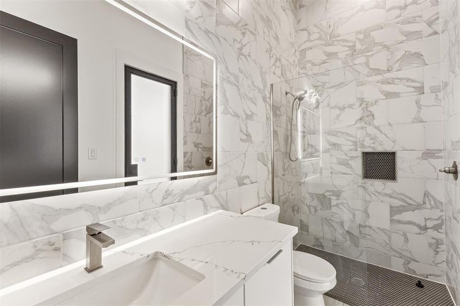 Modern bathroom featuring a white vanity with an integrated sink, a large illuminated mirror, and a walk-in shower with stone-look tile walls, a built-in niche, and dark penny tile flooring