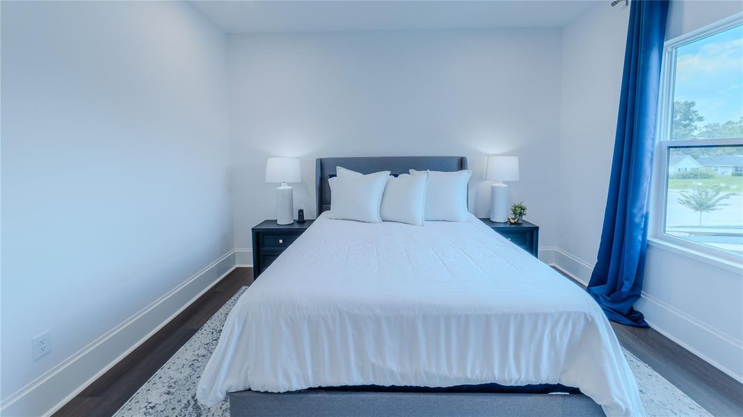 This bedroom features a cozy bed with white linens, flanked by two nightstands with lamps. Natural light pours in through a large window with blue curtains, creating a bright and inviting atmosphere.
