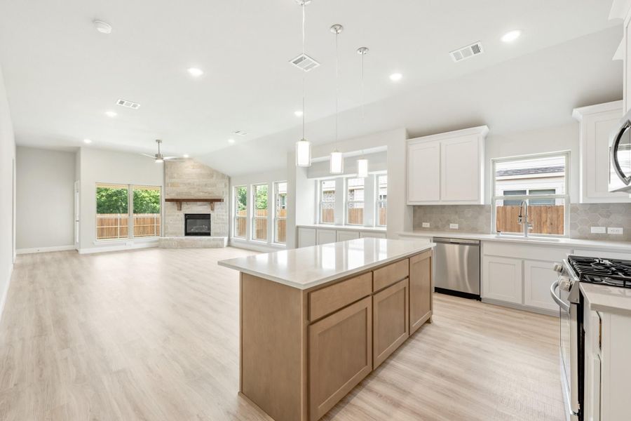 Open kitchen with center island, white cabinets, and light wood floors flowing into living room with stone fireplace. Open kitchen with center island, white cabinets, and light wood floors flowing into living room with stone fireplace.