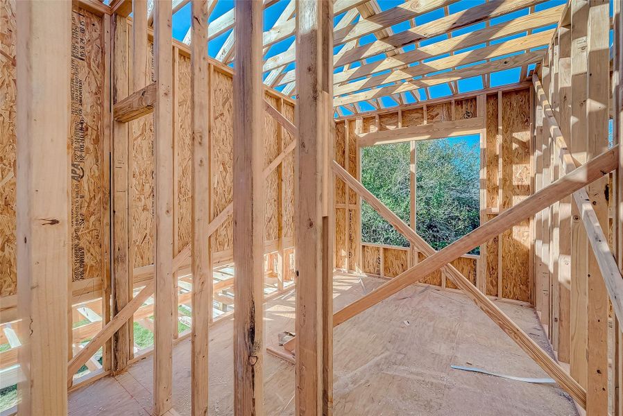 This photo shows the interior of a house under construction, featuring wooden framing and an unfinished roof. Natural light comes through a large window opening, highlighting the spacious layout.