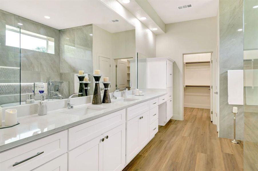 Spacious bathroom featuring a dual vanity with white cabinetry and light-toned countertops, wood-finish flooring, a large glass-enclosed shower with tile surround, and recessed lighting Spacious bathroom featuring a dual vanity with white cabinetry and light-toned countertops, wood-finish flooring, a large glass-enclosed shower with tile surround, and recessed lighting