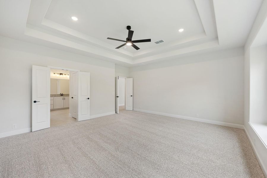Large bedroom with tray ceiling, ceiling fan, carpet floors, and open doors to bathroom and closet Large bedroom with tray ceiling, ceiling fan, carpet floors, and open doors to bathroom and closet
