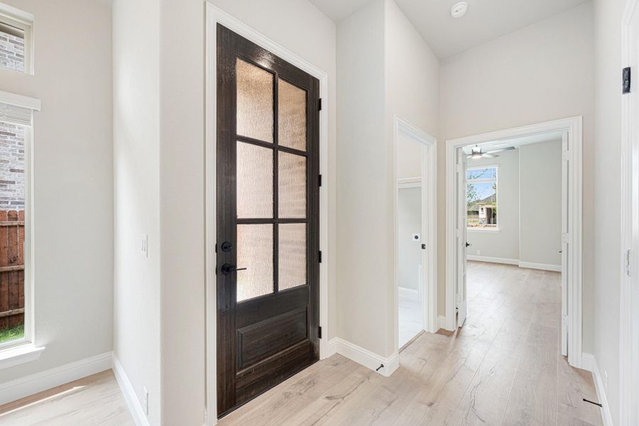 Hallway with dark wood glass-panel door, white walls, and light hardwood floors leading to a bedroom