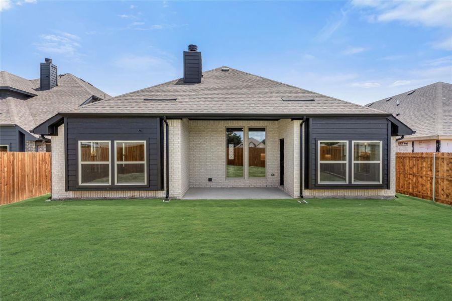 Rear exterior of single-story home with white brick, dark siding, covered patio, and fenced backyard with green lawn