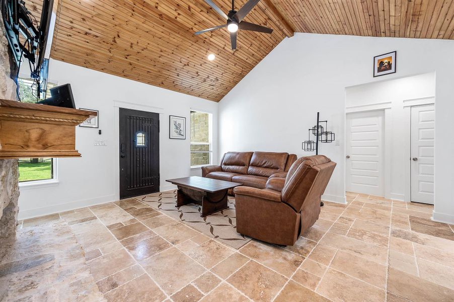 Natural light fills this cozy living room, highlighting the soaring wood-paneled ceiling, spacious layout, and inviting stone fireplace with a rustic wood mantel. The combination of warm finishes and comfortable design creates a relaxing everyday living space.