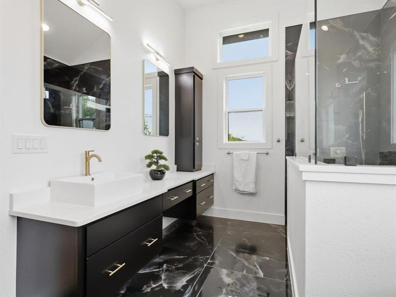 Spacious bathroom featuring black and white veined flooring, a dual vanity with white countertops, black cabinetry with gold-toned hardware, a vessel sink with a gold-toned faucet, and an enclosed shower with black and white veined wall tiling