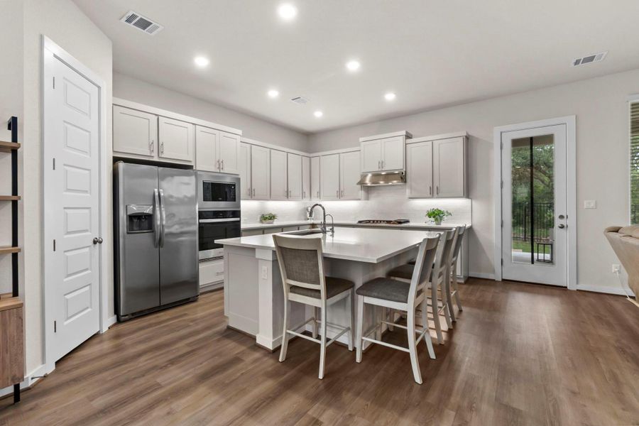 Open-concept kitchen featuring an oversized central island with seating, stainless steel appliances, light gray cabinetry, recessed lighting, and wood-finish flooring