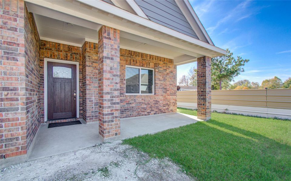This photo features a welcoming front porch with brick construction and a dark wooden door, surrounded by a small, neatly maintained lawn. The covered entryway provides a cozy outdoor space.