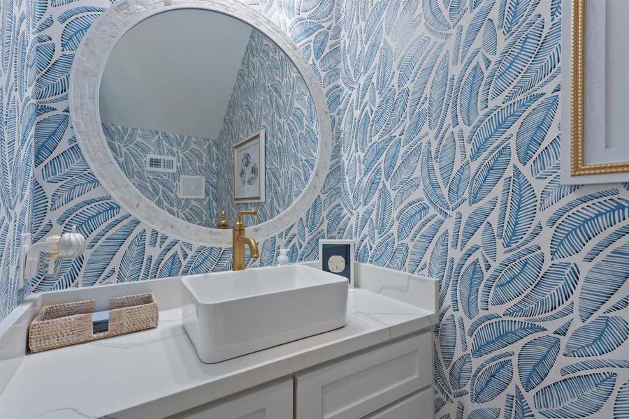 The main area of the fist floor offers a half bathroom with stylish Serena & Lily designer wallpaper. Blue palm leaf-patterned wallpaper and a modern white vanity with a vessel sink and brass faucet.