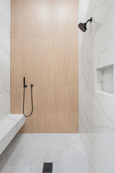 Spacious shower featuring white marble-patterned tile, wood-finish accent wall, built-in bench, recessed shelf, and matte black fixtures