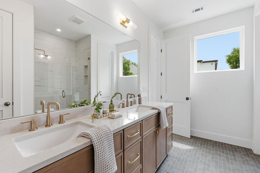 Bathroom featuring a dual vanity with light-toned countertops and wood-finish cabinetry, a large frameless mirror, and brushed gold fixtures