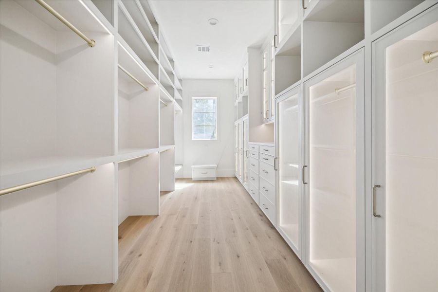 One of two well-lit walk-in closets featuring ample shelving, hanging rods, and cabinetry with a sleek, modern design. The light wood floors and the natural light from the window. One of two well-lit walk-in closets featuring ample shelving, hanging rods, and cabinetry with a sleek, modern design. The light wood floors and the natural light from the window.