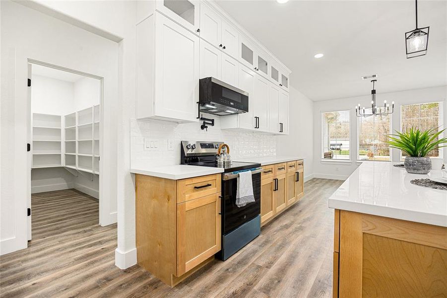 This modern kitchen features sleek white and wood cabinetry, a spacious island with quartz countertops, and stainless steel appliances. It includes a large walk-in pantry and is complemented by a bright dining area with large windows, offering plenty of natural light.