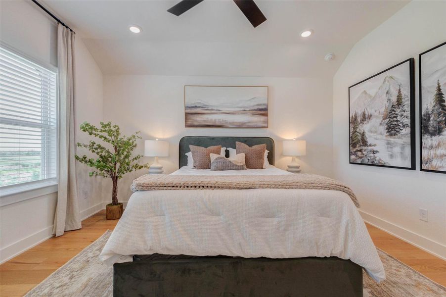 This inviting bedroom blends comfort and contemporary style with a plush upholstered bed, layered neutral bedding, and warm wood flooring. Natural light enters through the large window, while recessed lights and a sleek ceiling fan provide added brightness and comfort. Oversized landscape artwork and coordinated bedside lamps create a calm, balanced atmosphere ideal for relaxation.