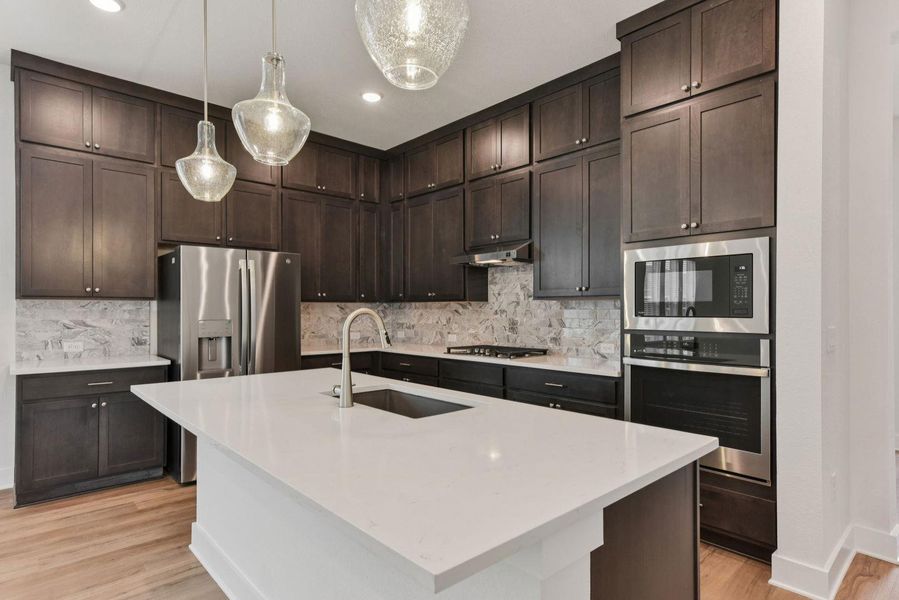 Kitchen featuring a center island with sink, dark wood cabinetry, stainless steel appliances, a stone tile backsplash, and light wood-finish flooring