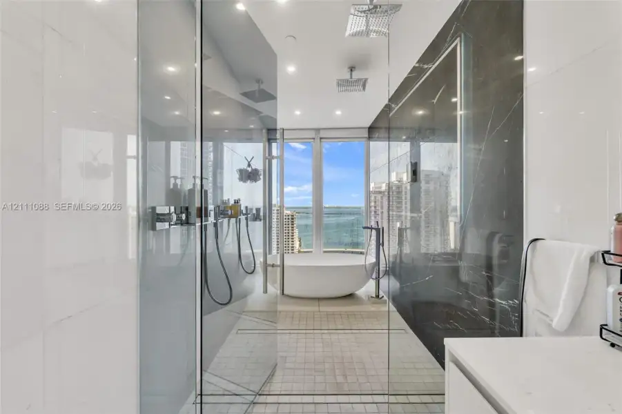 Walk-in glass-enclosed shower featuring dual rain-style showerheads and handheld fixtures, freestanding soaking tub, contrasting large-format wall tiling, and floor-to-ceiling windows Walk-in glass-enclosed shower featuring dual rain-style showerheads and handheld fixtures, freestanding soaking tub, contrasting large-format wall tiling, and floor-to-ceiling windows