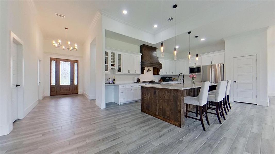 Open-concept living area featuring light wood-finish flooring, a gourmet kitchen with a wood-wrapped island, stainless steel appliances, and white cabinetry, complemented by a prominent wood-finished range hood and a chandelier in the entryway Open-concept living area featuring light wood-finish flooring, a gourmet kitchen with a wood-wrapped island, stainless steel appliances, and white cabinetry, complemented by a prominent wood-finished range hood and a chandelier in the entryway