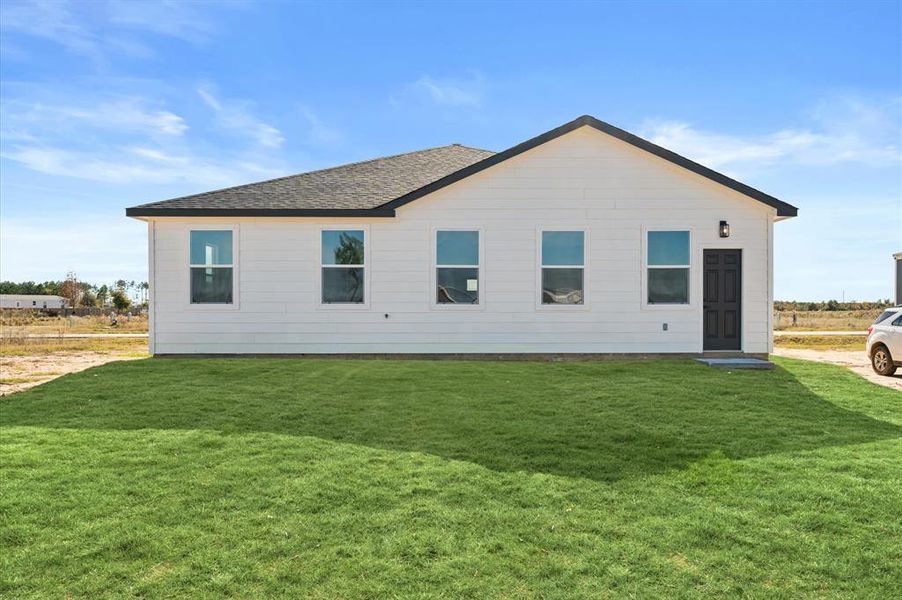 This is a modern single-story house featuring a clean, white exterior with a dark roof and trim. The home has several windows offering natural light, a neatly manicured lawn, and a simple, welcoming entryway. It appears to be in a new or developing neighborhood.