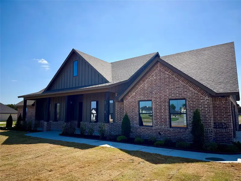 View of front of home featuring covered porch, a front lawn, board and batten siding, a shingled roof, and brick siding View of front of home featuring covered porch, a front lawn, board and batten siding, a shingled roof, and brick siding