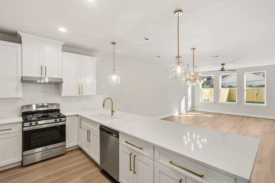 This photo showcases a modern open-concept kitchen and living area. The kitchen features sleek white cabinetry, a stainless steel stove, and a dishwasher, complemented by elegant pendant lighting. The spacious living area has large windows that provide ample natural light and views of a fenced backyard.