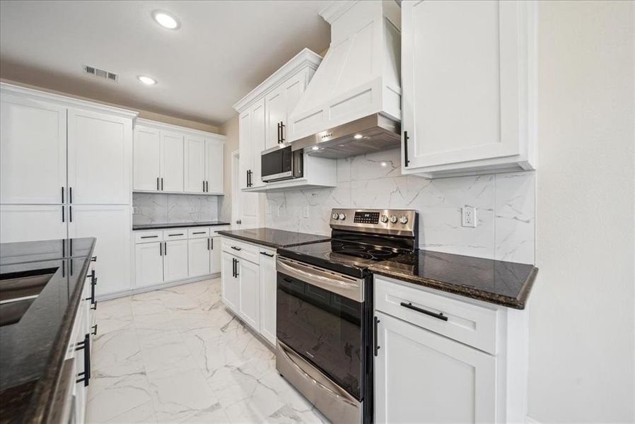 Kitchen with appliances with stainless steel finishes, premium range hood, tasteful backsplash, dark stone counters, and white cabinetry