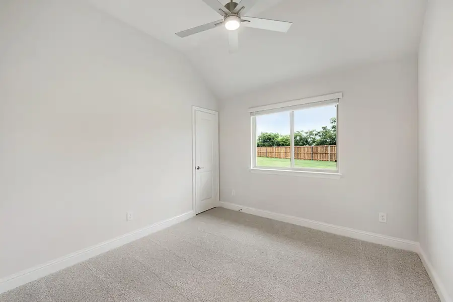 Empty bedroom with vaulted ceiling, ceiling fan, carpet flooring, and a window overlooking a fenced backyard Empty bedroom with vaulted ceiling, ceiling fan, carpet flooring, and a window overlooking a fenced backyard