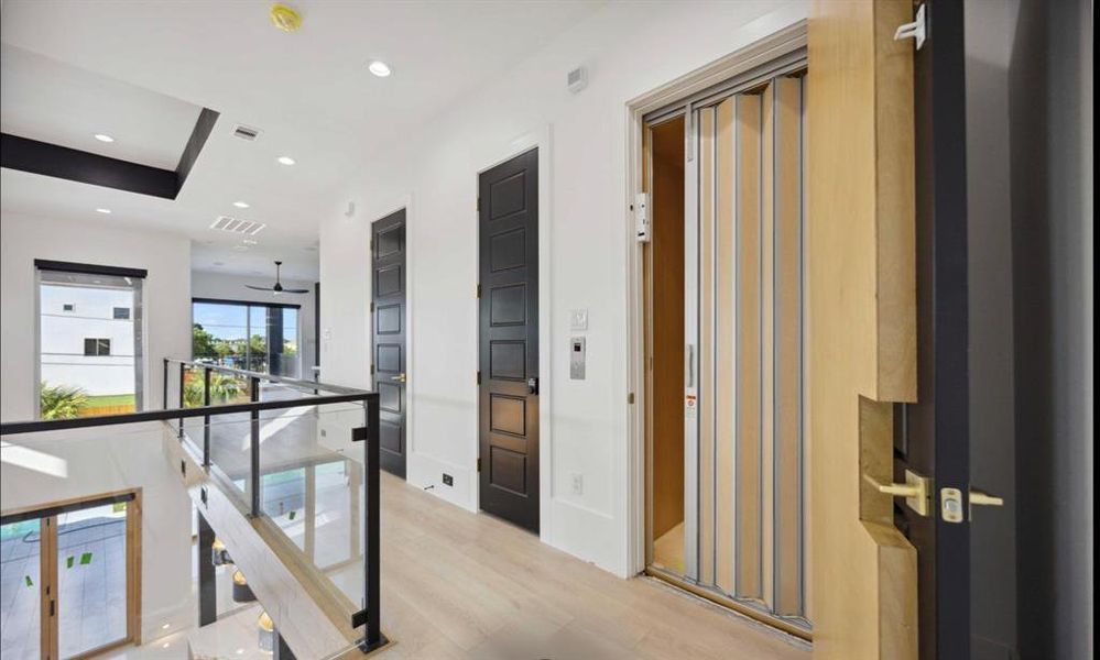 This is a modern, bright hallway featuring entry to 2 floor elevator door, light hardwood flooring, and a glass balustrade overlooking the lower level. It's well-lit with large windows and high ceilings, offering a spacious and airy feel.