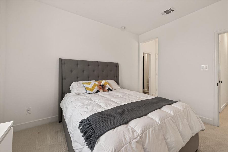 This photo showcases a cozy bedroom with neutral walls and carpet, featuring a comfortable bed with a tufted headboard. The room has a clean, minimalist design and includes an en-suite bathroom access.