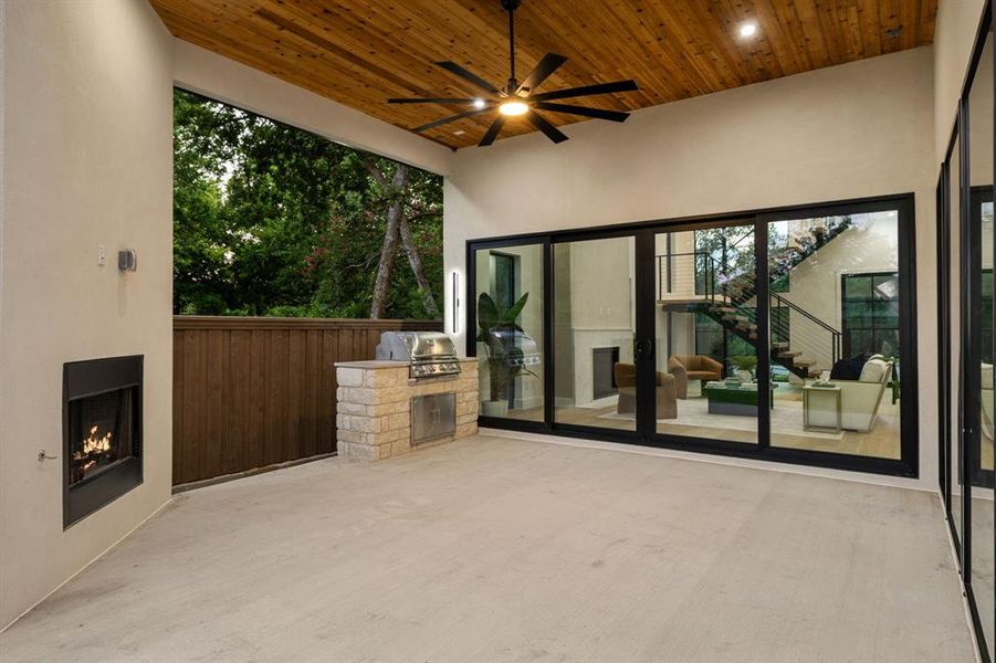 Covered patio featuring wood-paneled ceiling, ceiling fan, built-in fireplace, and stone-clad outdoor kitchen with integrated grill