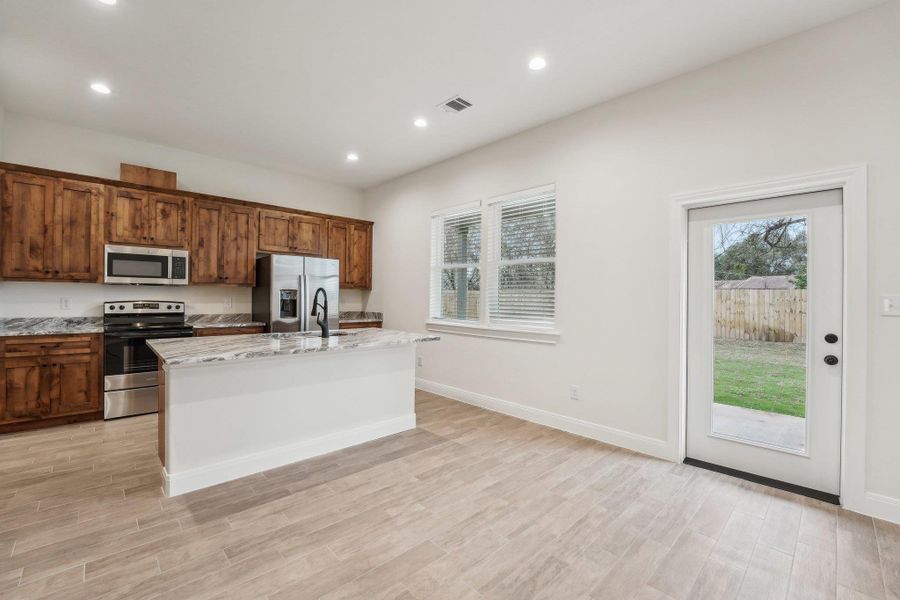 Kitchen with visible vents, a center island with sink, wood plank tile floor, stainless steel appliances, and light stone countertops Kitchen with visible vents, a center island with sink, wood plank tile floor, stainless steel appliances, and light stone countertops