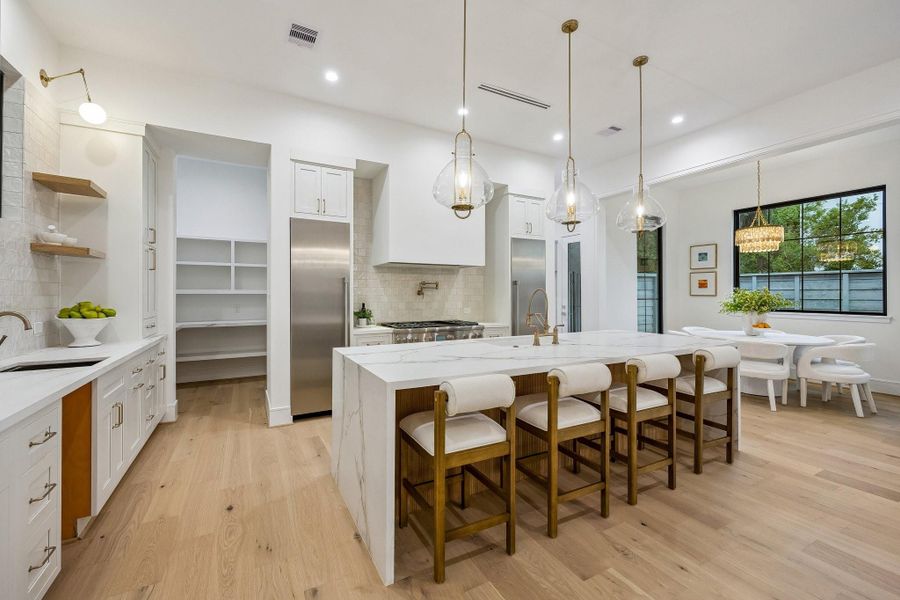 This kitchen has a bright, upscale contemporary design with warm touches and a soft, luxurious palette. The space feels open and airy, balancing clean architectural lines with rich textures. The large waterfall-edge island clad in dramatic white marble with subtle gray veining cascading down the sides. Three large glass pendant lights hang above the island, becoming sculptural focal points while keeping sightlines open because of their transparent design. Thermador Appliances, Inset Cabinetry & More...