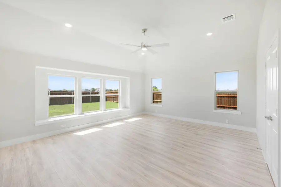 Empty room with light wood floors, ceiling fan, and large windows overlooking a fenced backyard